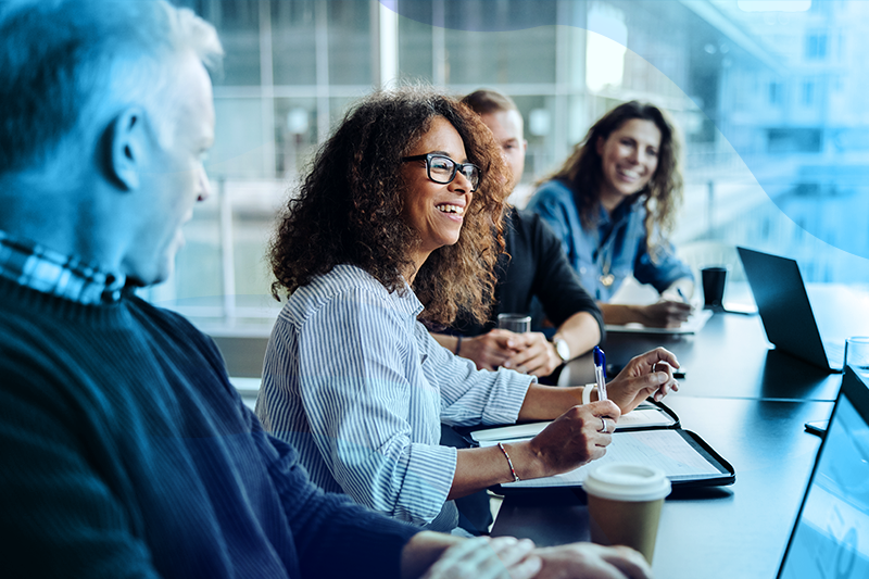 Group Of People Around A Conference Table Talking With Woman In The Middle Smiling In Conversation