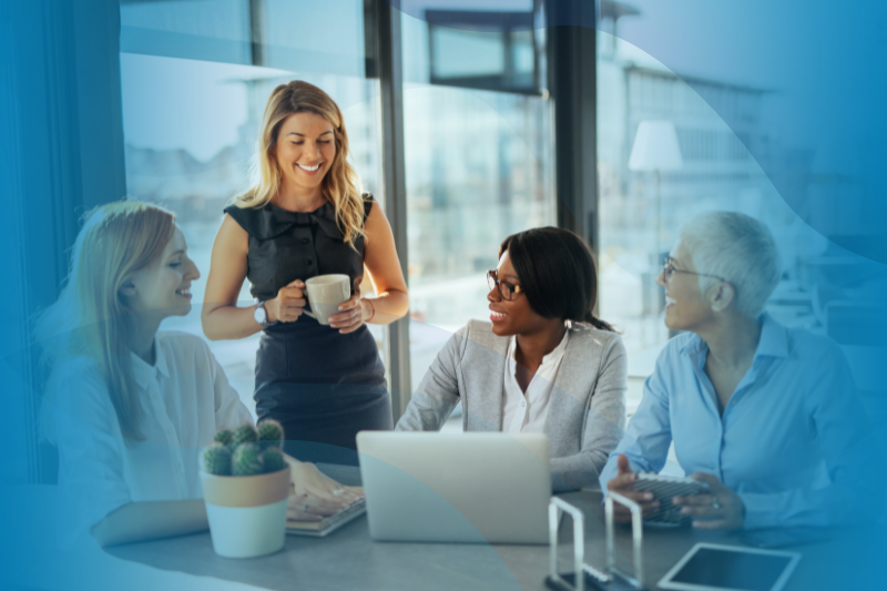 Group Of Women Talking At A Desk With One Standing Holding A Mug