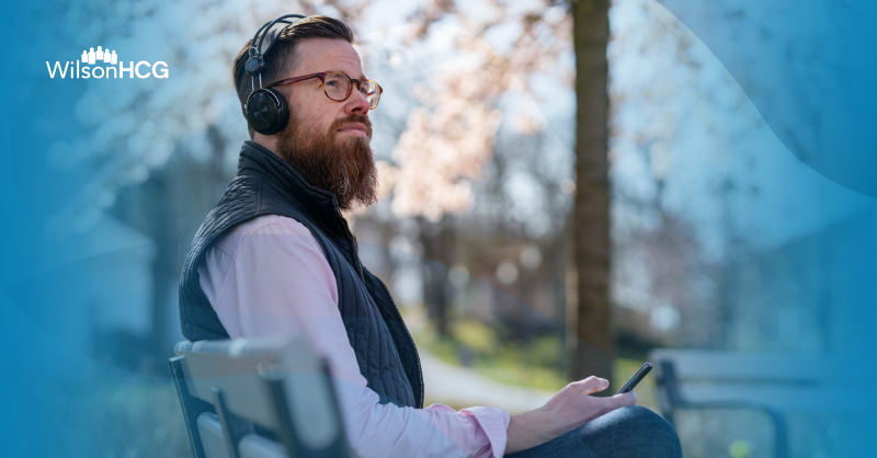 Man with glasses and beard with over-ear headphones listening to a podcast outside while sitting on a bench