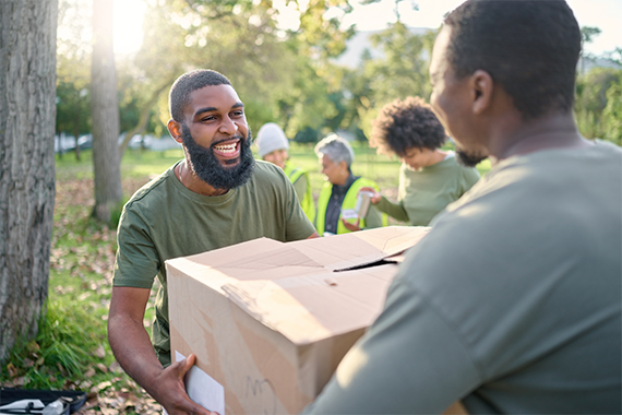 Two Black men carrying a big cardboard box outside doing group community work with others