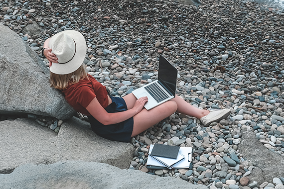 Woman working remotely on her laptop while sitting on rocks by a beach